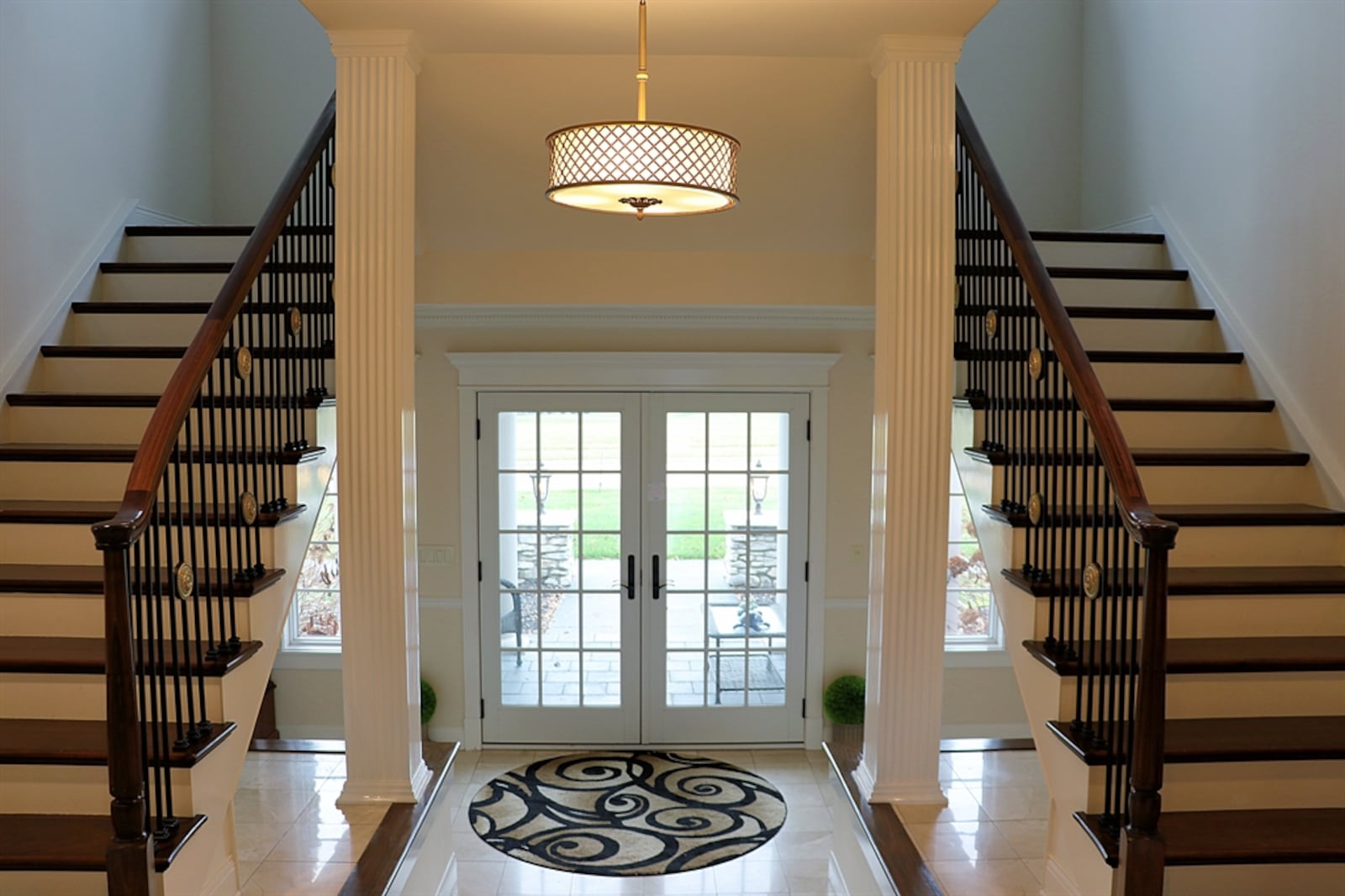 Double glass doors open into a grand foyer with polished ceramic-tile flooring and dark wood trim. Fluted columns accent the three-step entry to the main living space, and ceramic-tile display nooks are nestled under the split staircase.