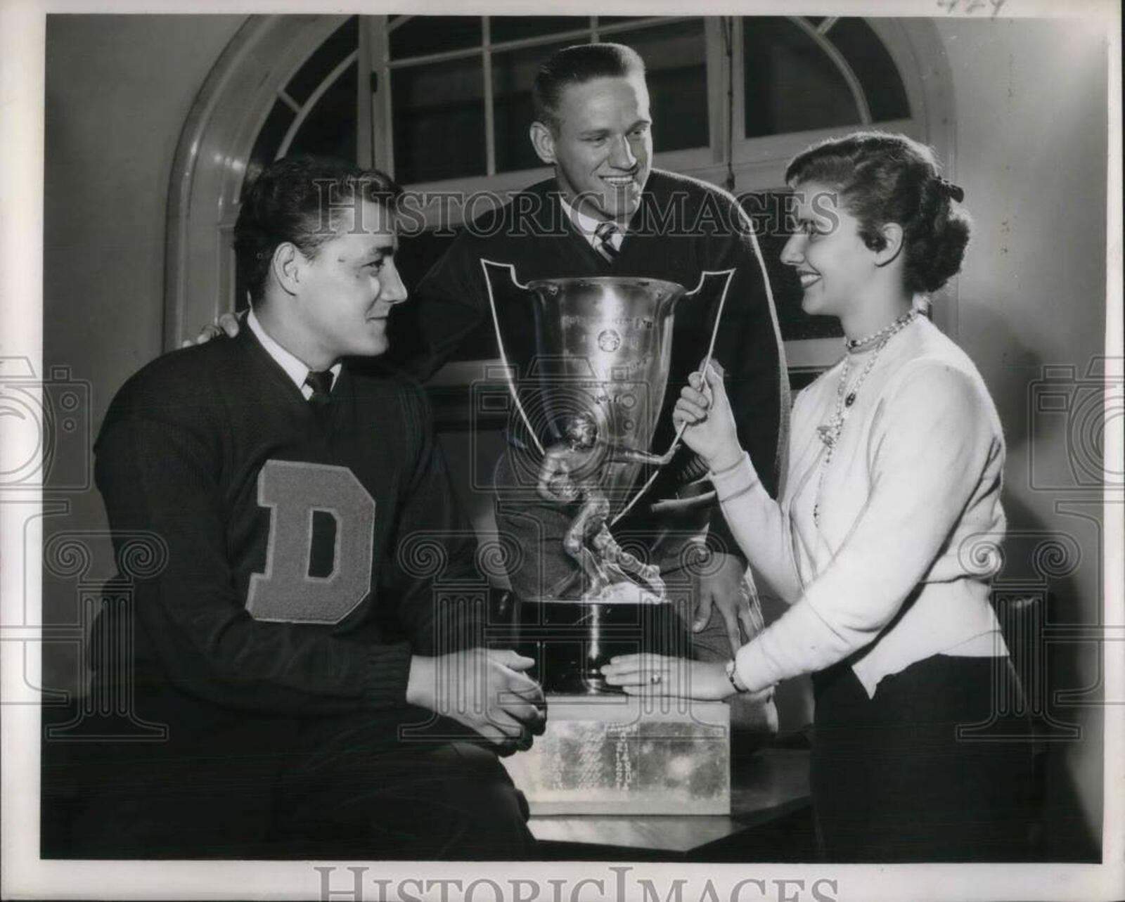 In October of 1954, Dayton Flyers standout Jim Katcavage is flanked by UD teammate Jack Muldowny (junior tackle) and Marilyn Stitch (freshman cheerleader) as they admire the Ohio Governor's Cup that Dayton captured with a victory over Xavier. The Cup tradition had begin in 1929. University of Dayton photo