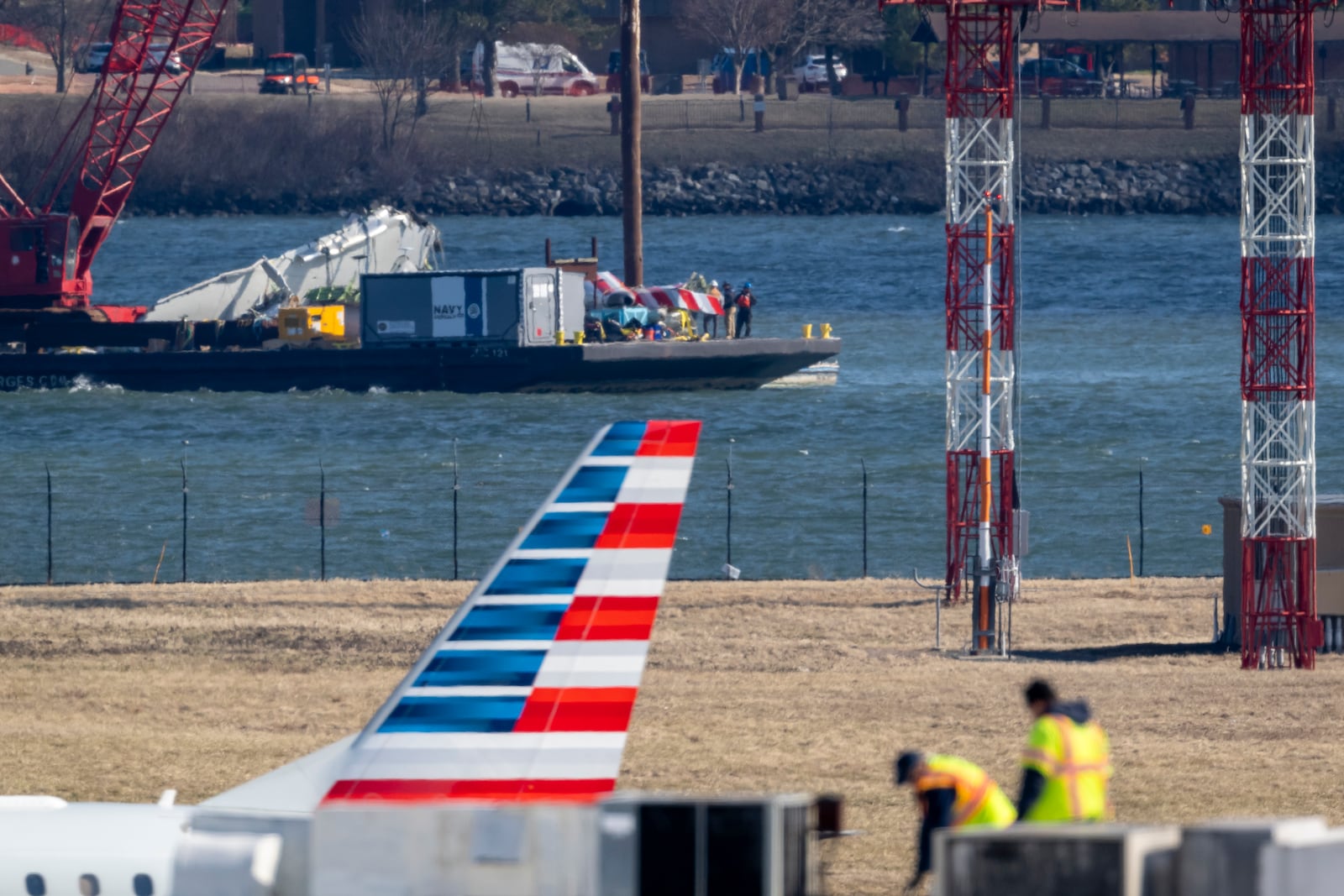 Parts of a plane are lifted from the water near the wreckage site in the Potomac River of a mid-air collision between an American Airlines jet and a Black Hawk helicopter, at Ronald Reagan Washington National Airport, Tuesday, Feb. 4, 2025, in Arlington, Va., as an American Airlines aircraft is seen landing on the runway, foreground. (AP Photo/Ben Curtis)