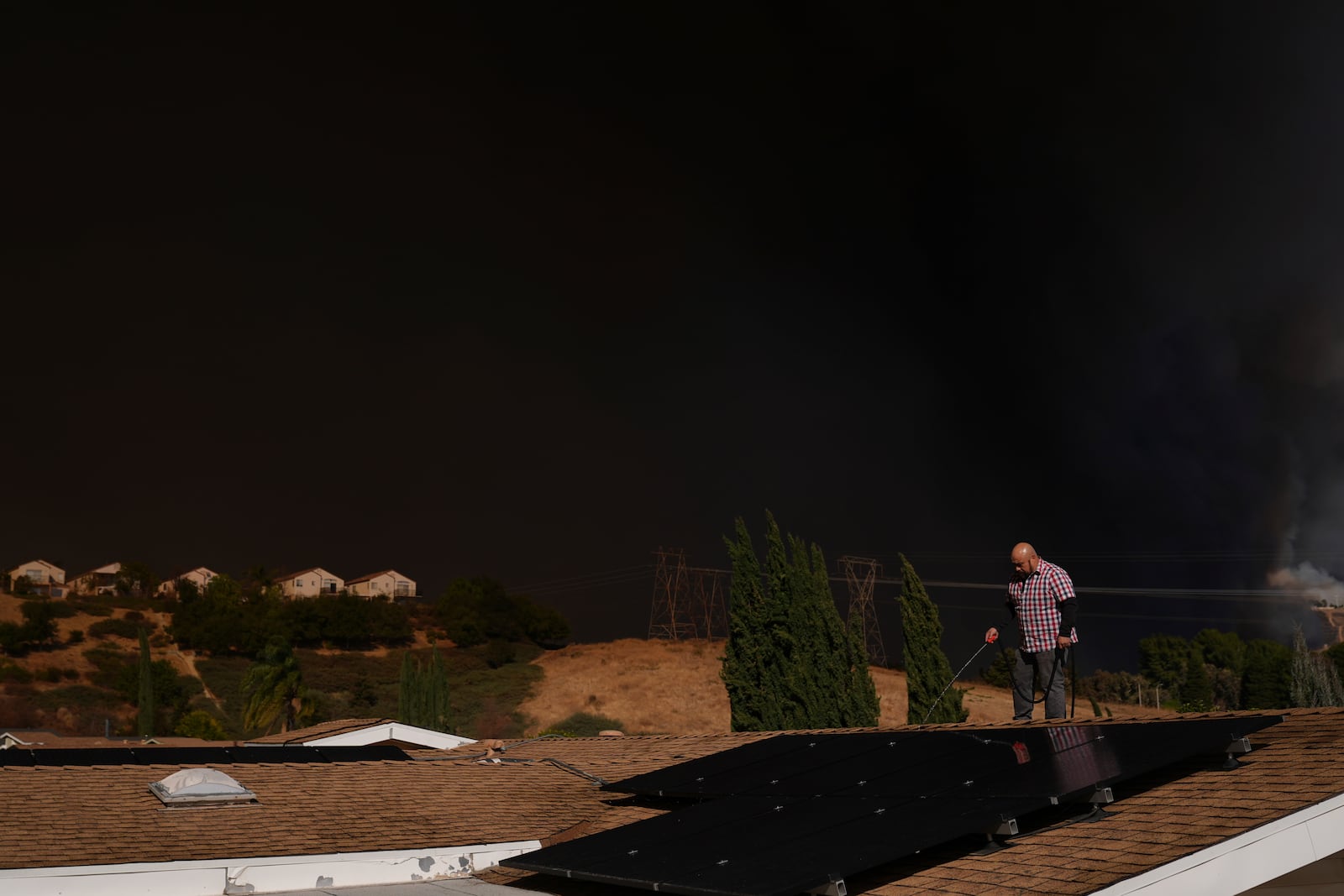 A home owner sprays water from the top of the roof at his brother's home Castaic, Calif., as a large plume of smoke caused by the Hughes Fire rises from Castaic Lake Wednesday, Jan. 22, 2025. (AP Photo/Marcio Jose Sanchez)