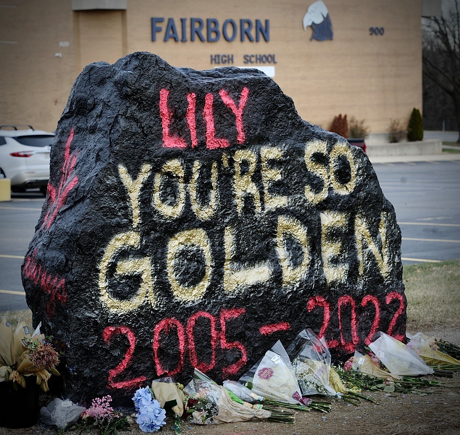 A large rock painted with "Lily, you're so golden, 2005-2022" in red and yellow spraypaint, with a memorial of flowers surrounding the base.