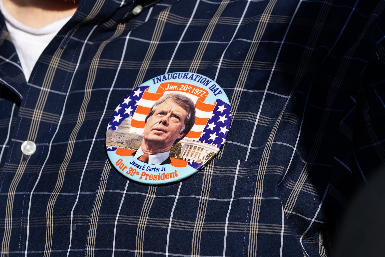 A person wears an inauguration button for  former President Jimmy Carter as his funeral procession moves through Plains, Ga., Saturday, Jan. 4, 2025. (Dustin Chambers/The New York Times)