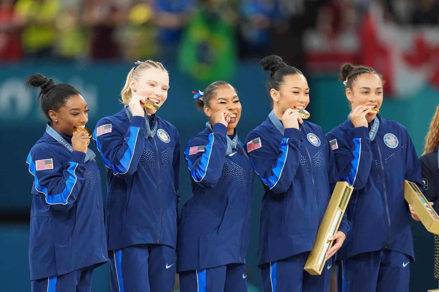 
                        Fom left: Simone Biles, Jade Carey, Jordan Chiles, Sunisa Lee and Hezly Rivera of the U.S. bite their gold medals at the medal ceremony in the women's artistic gymnastics team finals at the 2024 Summer Olympics at Bercy Arena in Paris, on Tuesday, July 30, 2024. (Chang W. Lee/The New York Times)
                      