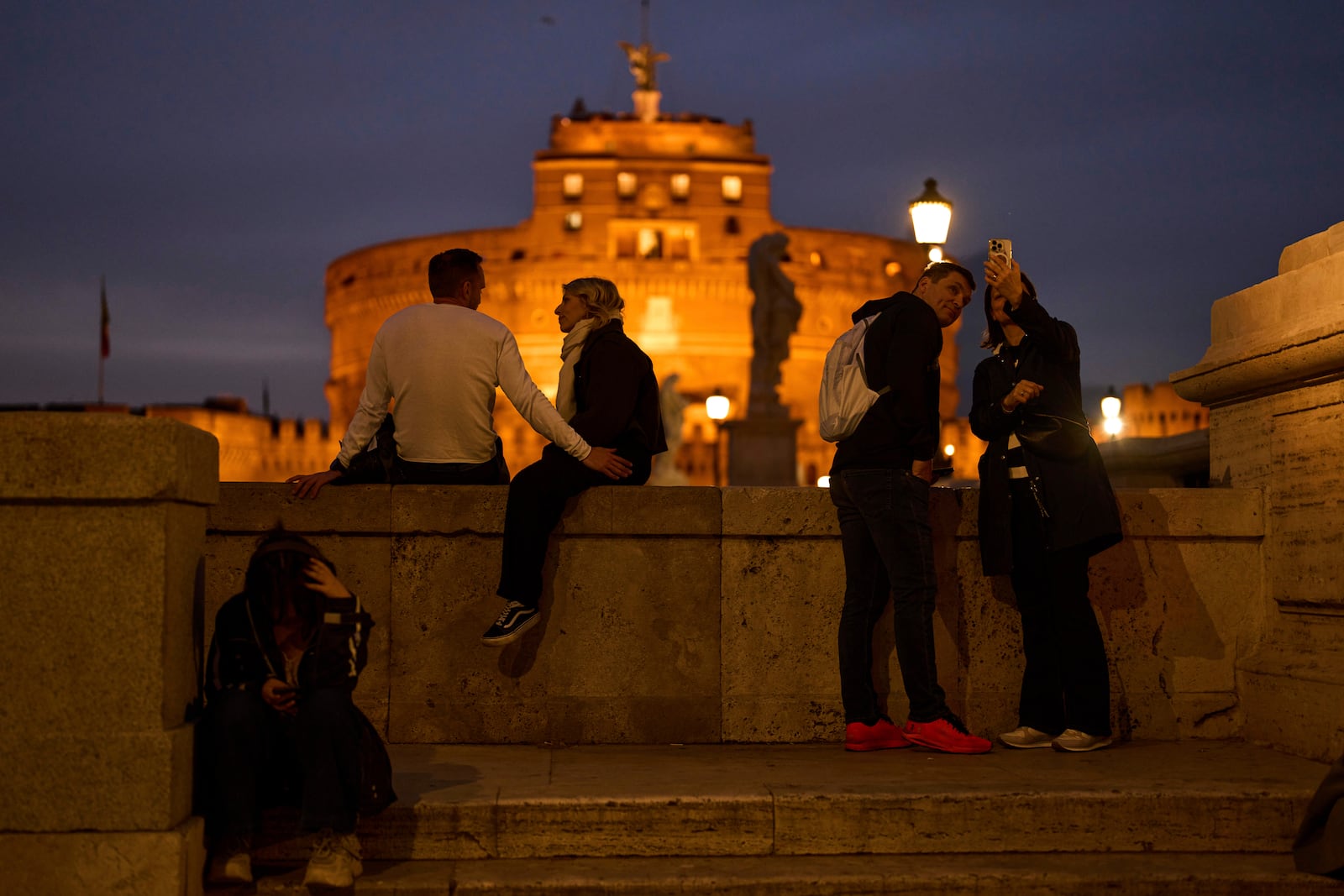 Tourists sit near Sant'Angelo castle, background, as the sun sets in Rome, Italy, Friday, March 7, 2025. (AP Photo/Francisco Seco)