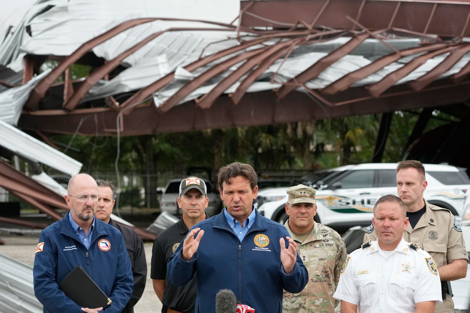 Florida Gov. Ron DeSantis, center, speaks during a news conference in front of a St. Lucie County Sheriff's parking facility that was damaged by a tornado spawned ahead of Hurricane Milton destroyed it, Thursday, Oct. 10, 2024, in Fort Pierce. (AP Photo/Wilfredo Lee)