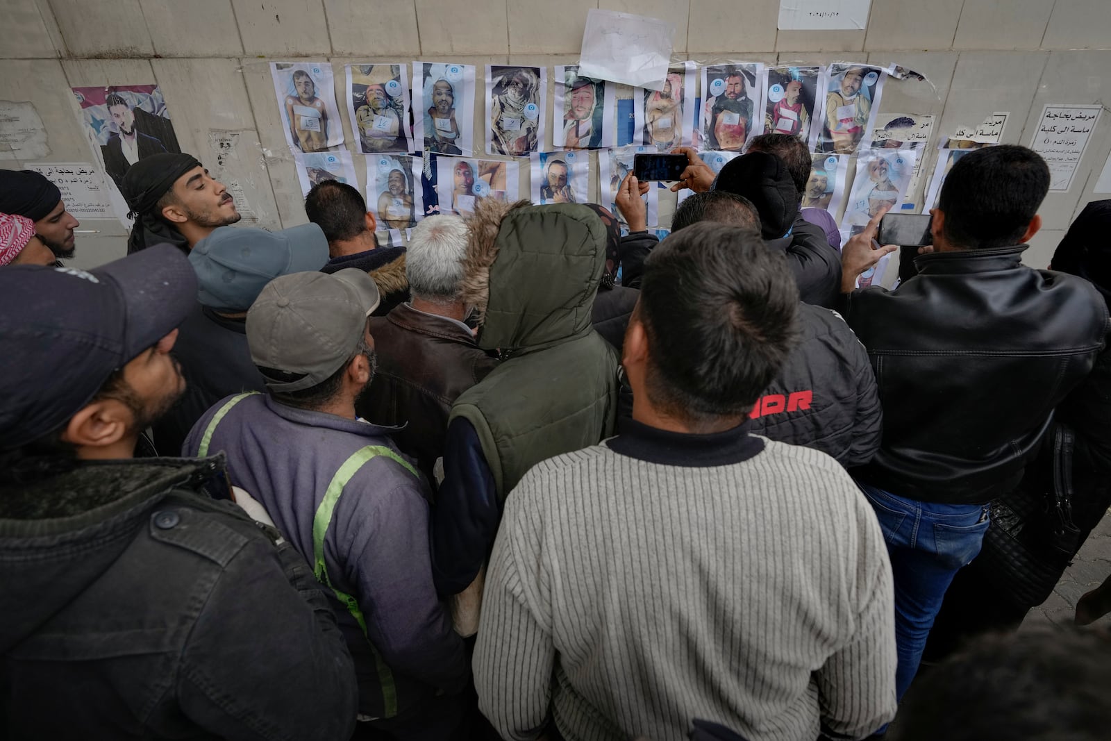 People examine photos as they search for missing or deceased people on the wall outside the morgue of Al-Mojtahed Hospital in Damascus, Syria, on Wednesday, Dec. 11, 2024. Many are flocking to morgues, hoping to identify loved ones who have been killed or were imprisoned under the rule of President Bashar Assad, whose government collapsed over the weekend. (AP Photo/Hussein Malla)