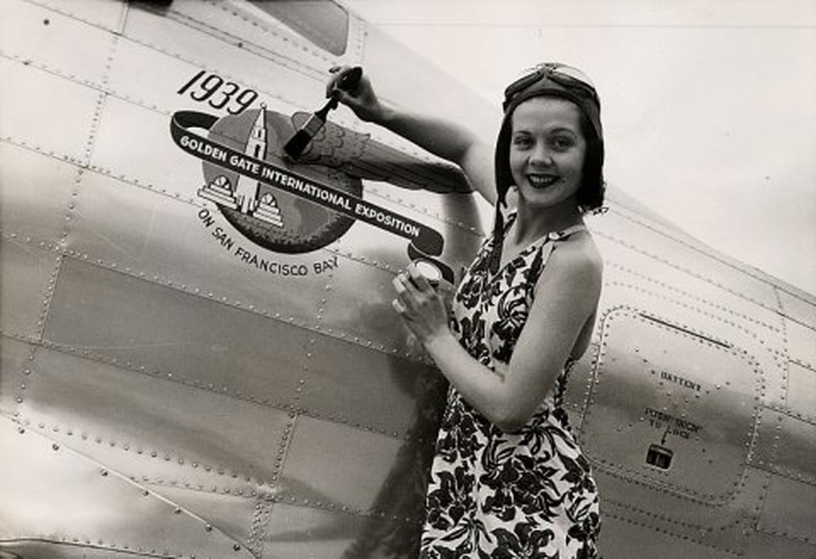 Captioned Zoe Dell Lantis, “most photographed girl in the world,” puts finishing touches to 1939 Golden Gate International Exposition emblem on the plane of Frank W. Fuller, Jr., air speed king named Chief Pilot of the Exposition. (Photo: San Francisco History Center, San Francisco Public Library via ia Engineers Club of Dayton)