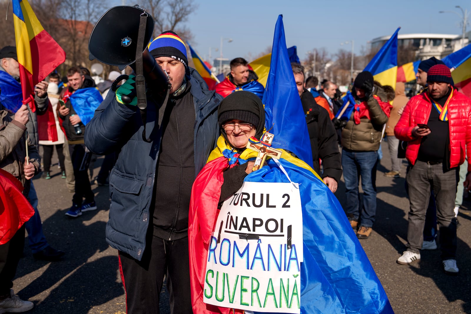 A woman draped in the Romanian flag sits next to a man shouting slogans during a protest by supporters of Calin Georgescu, the winner of Romania's first round of presidential election which the Constitutional Court later annulled in Bucharest, Romania, Monday, Feb. 10, 2025. (AP Photo/Vadim Ghirda)