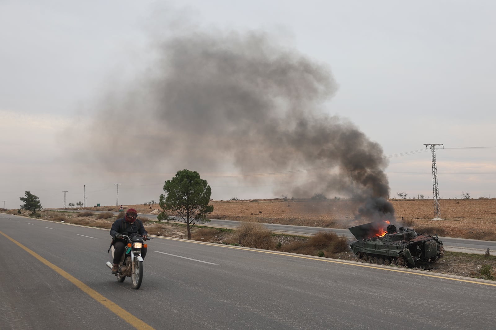 A man rides his motorcycle past a burning government armoured vehicle south of Hama, Syria, Saturday, Dec. 7, 2024. (AP Photo/Ghaith Alsayed)