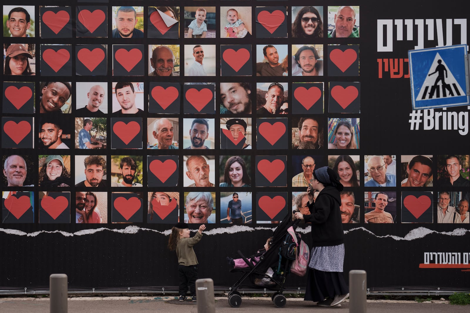 FILE - A woman and her children walk past a wall with photographs of hostages who were kidnapped during the Oct. 7, 2023, Hamas cross-border attack in Israel, Feb. 26, 2024, in Jerusalem. On Monday, June 24, Israelis who were taken hostage or lost loved ones during the attack filed suit against the United Nations agency that aids Palestinians, claiming it has helped finance the militants by paying agency staffers in U.S. dollars and thereby funneling them to money-changers in Gaza who allegedly give a cut to Hamas. (AP Photo/Leo Correa, File)