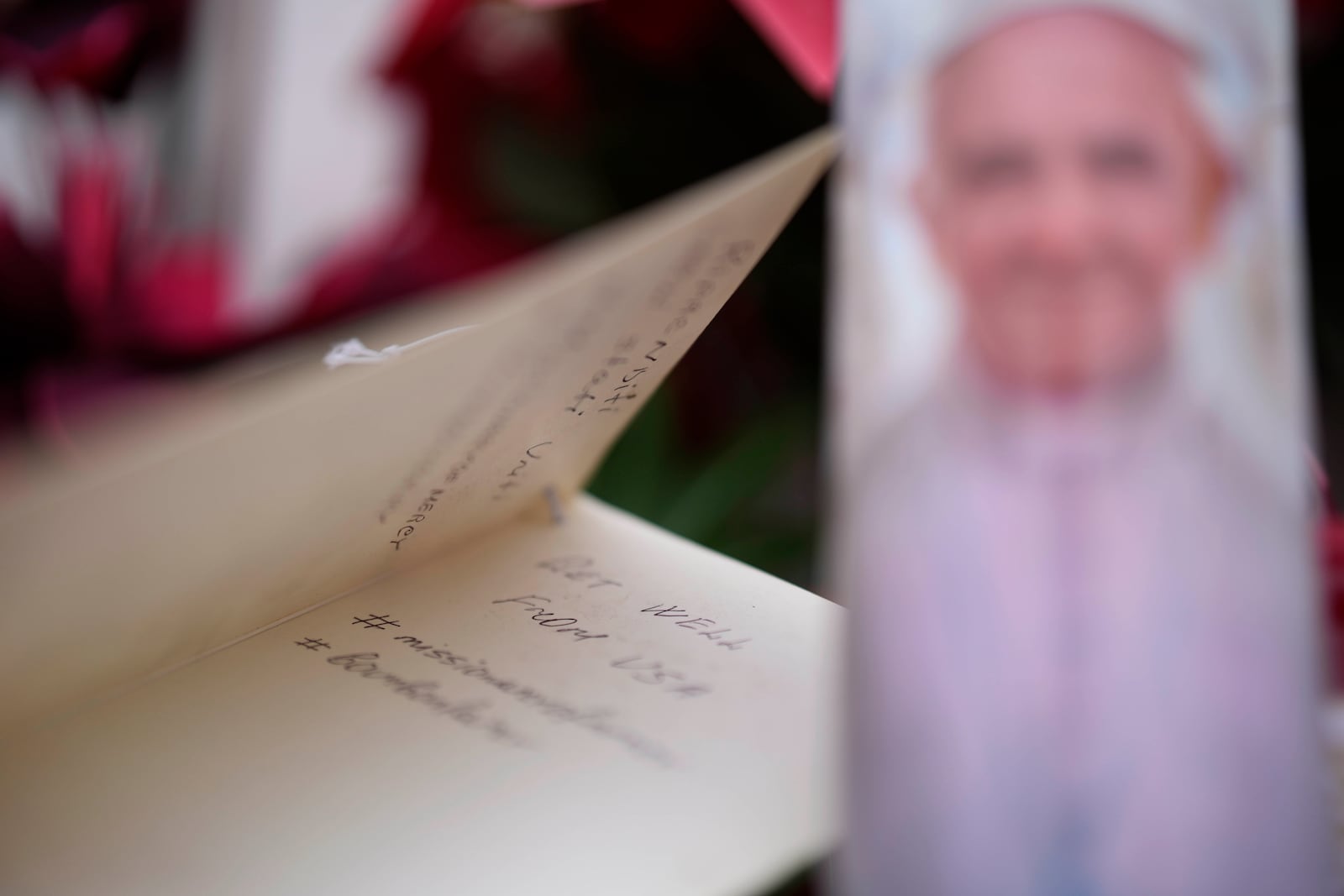 A well wishing card lies near candles adorned with pictures of Pope Francis laid under the statue of late Pope John Paul II outside Agostino Gemelli Polyclinic in Rome, Wednesday, Feb. 19, 2025, where the Pontiff is hospitalized since Friday, Feb. 14. (AP Photo/Gregorio Borgia)