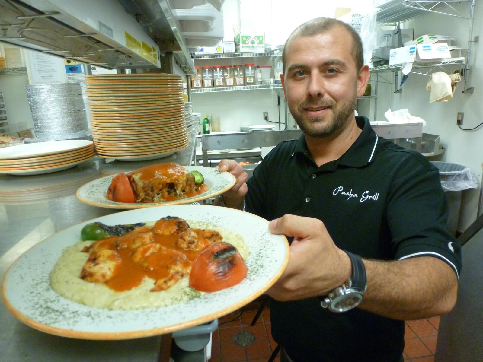 J. Erme Bektas, executive chef and owner of Pasha Grill in Beavercreek holds a plate of smoked eggplant puree topped with grilled chicken breast and fresh tomato sauce in the foreground and minced beef and lamb layered between slices of eggplant and topped with sauce and cheese. Contributed photo by Alexis Larsen