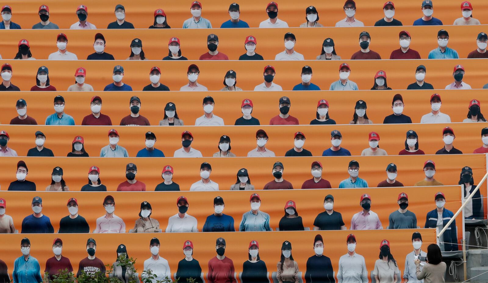 A woman holds her smartphone in front of the spectators' seats which are covered with pictures of fans, before the start of a baseball game between Hanwha Eagles and SK Wyverns in Incheon, South Korea, Tuesday, May 5, 2020. Cheerleaders danced beneath rows of empty seats and umpires wore protective masks as a new baseball season began in South Korea. After a weeks-long delay because of the coronavirus pandemic, a hushed atmosphere allowed for sounds like the ball hitting the catcher's mitt and bats smacking the ball for a single or double to echo around the stadium. (AP Photo/Lee Jin-man)