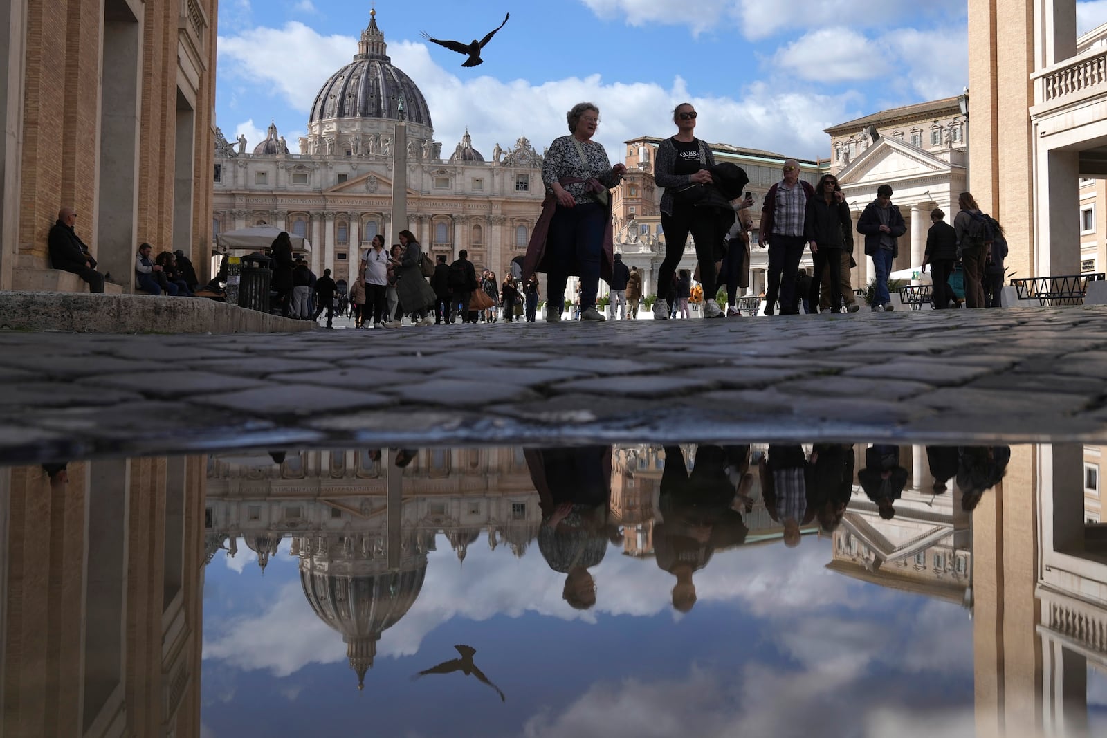 People are reflected in a puddle as they walk in in front of St. Peter's Square at the Vatican, Thursday, Feb. 27, 2025. (AP Photo/Alessandra Tarantino)