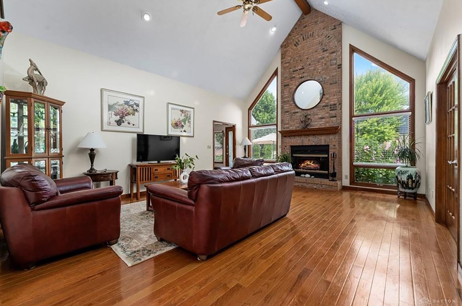 The two story great room features a brick fireplace, beamed ceiling, wood flooring and windows surrounding the two story brick gas fireplace.