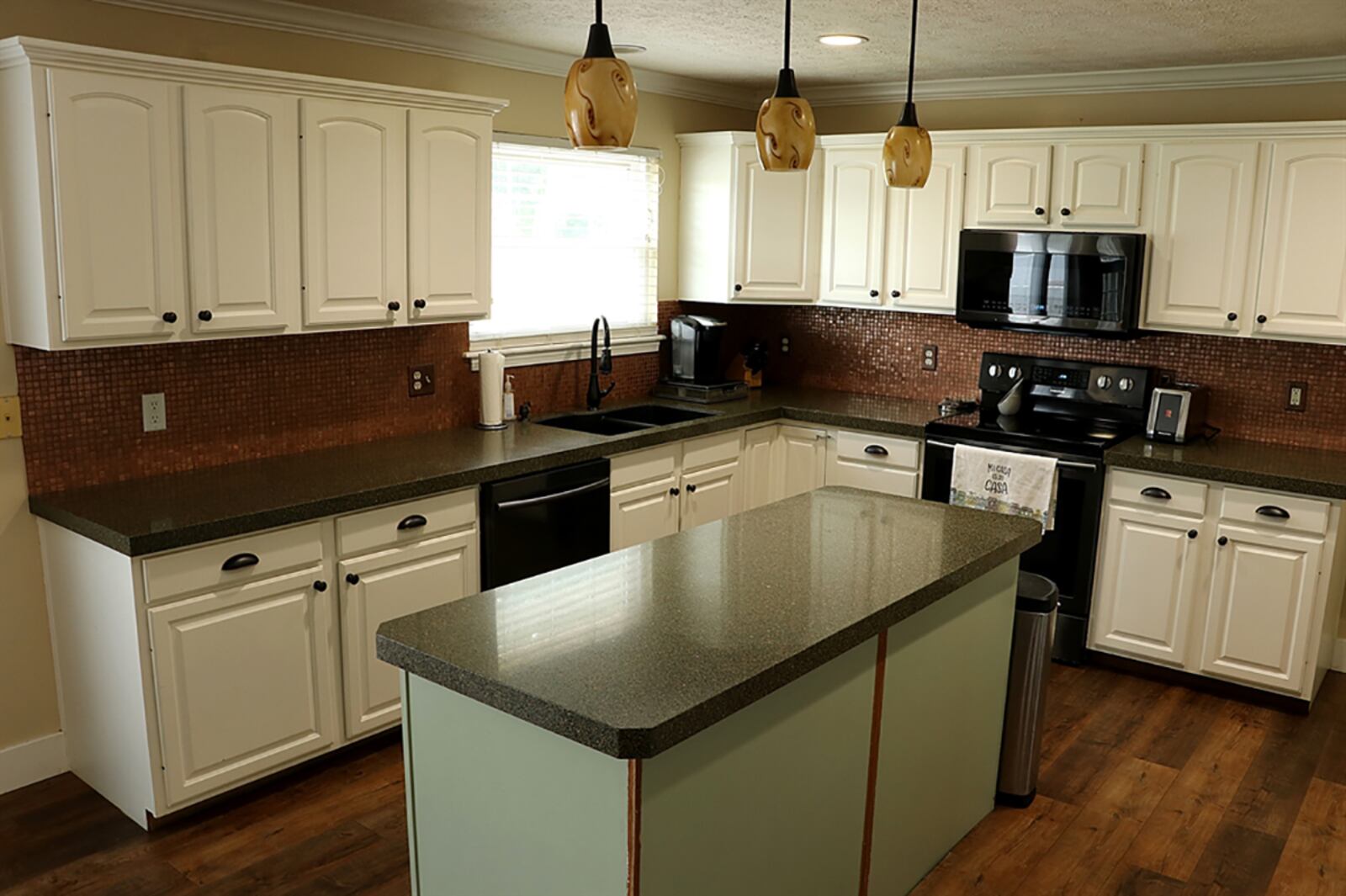 A large kitchen island offers storage and breakfast bar space. Teardrop light features hang above the island. White cabinetry is complemented by dark countertops and copper mosaic tiles accent the wall space. CONTRIBUTED PHOTO BY KATHY TYLER