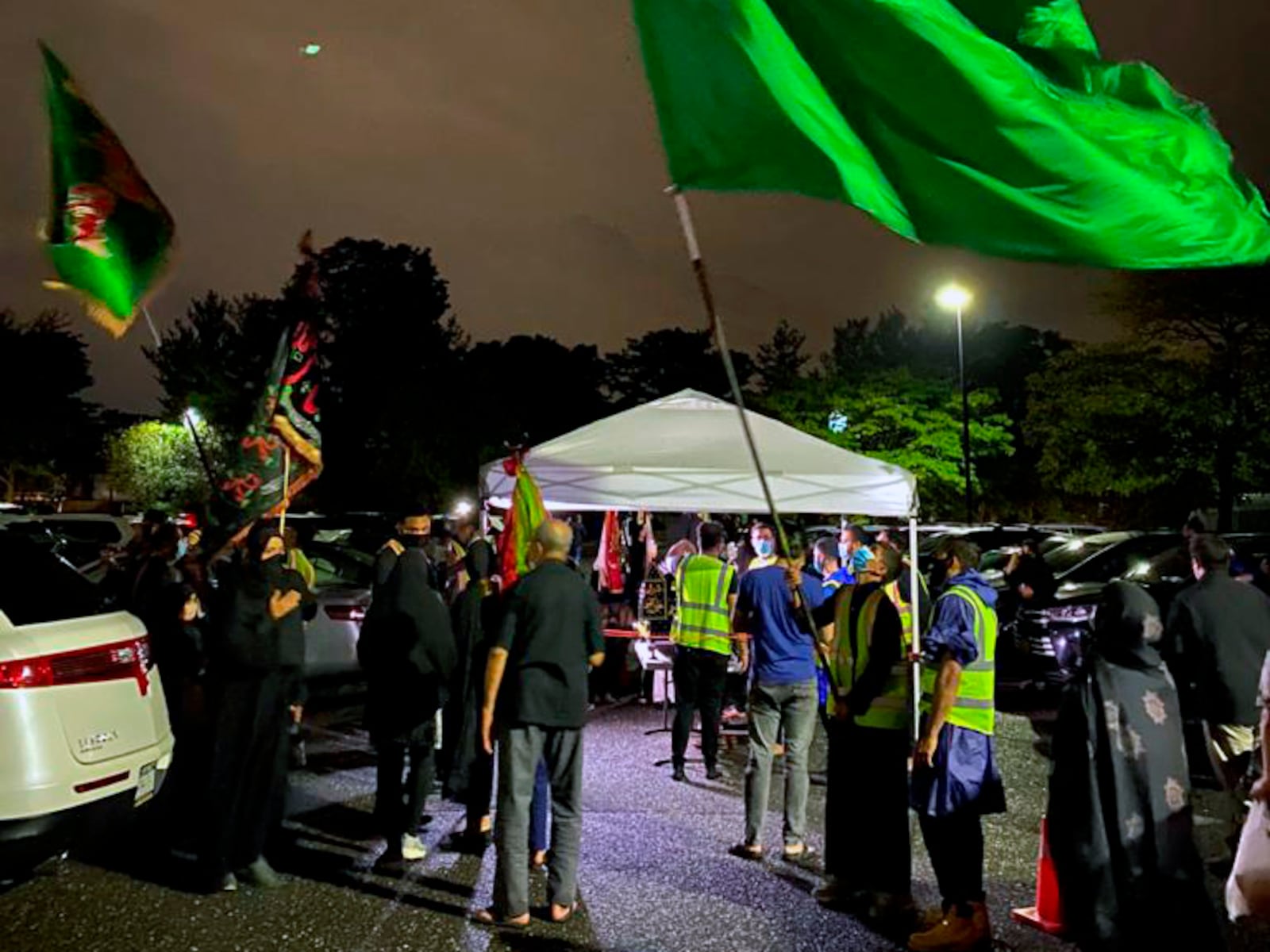 In a photo provided by Fatima Mukhi-Siwji, people gather with flags in a parking lot for a drive-in commemoration of the seventh-century death of Imam Hussein, Thursday, Aug. 27, 2020, in Hicksville, N.Y.  Shiite Muslims are marking the mourning period in the shadow of the coronavirus. (Fatima Mukhi-Siwji via AP)(Fatima Mukhi-Siwji via AP)