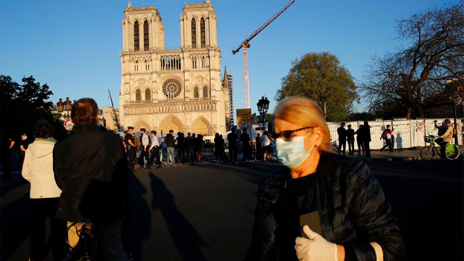 A woman wearing a mask to protect against coronavirus walks past people stopped in front of Notre Dame Cathedral as the bells ring to mark one year of blaze Wednesday, April 15, 2020 in Paris. A year after a devastating fire, the restoration of Notre Dame Cathedral has been halted by a lockdown in Paris to battle the coronavirus. The April 15, 2019, blaze gutted its interior, toppled its famous spire and horrified the world.