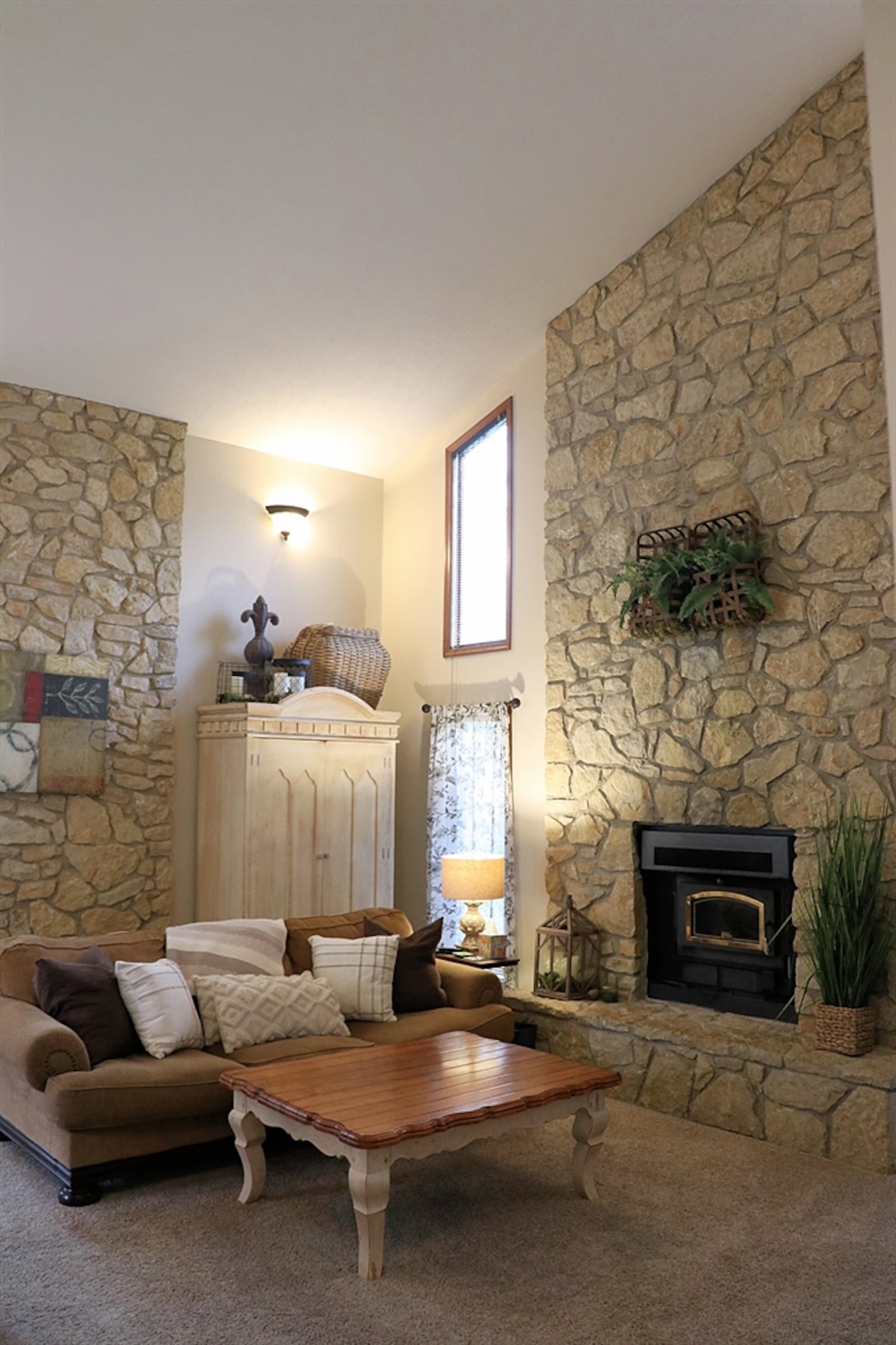 Stone accents two walls of the great room, which features a two-story fireplace with an elevated stone hearth. Tall windows flank the fireplace. 