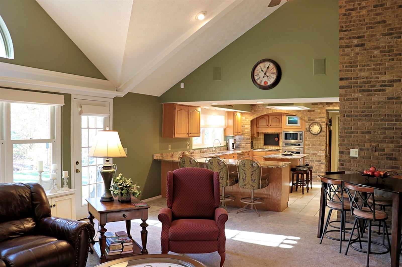 Great room has a vaulted ceiling, a corner brick wood-burning fireplace, and a wall of windows with built-in window seats and cabinetry. A peninsula counter divides the kitchen from the great room. 