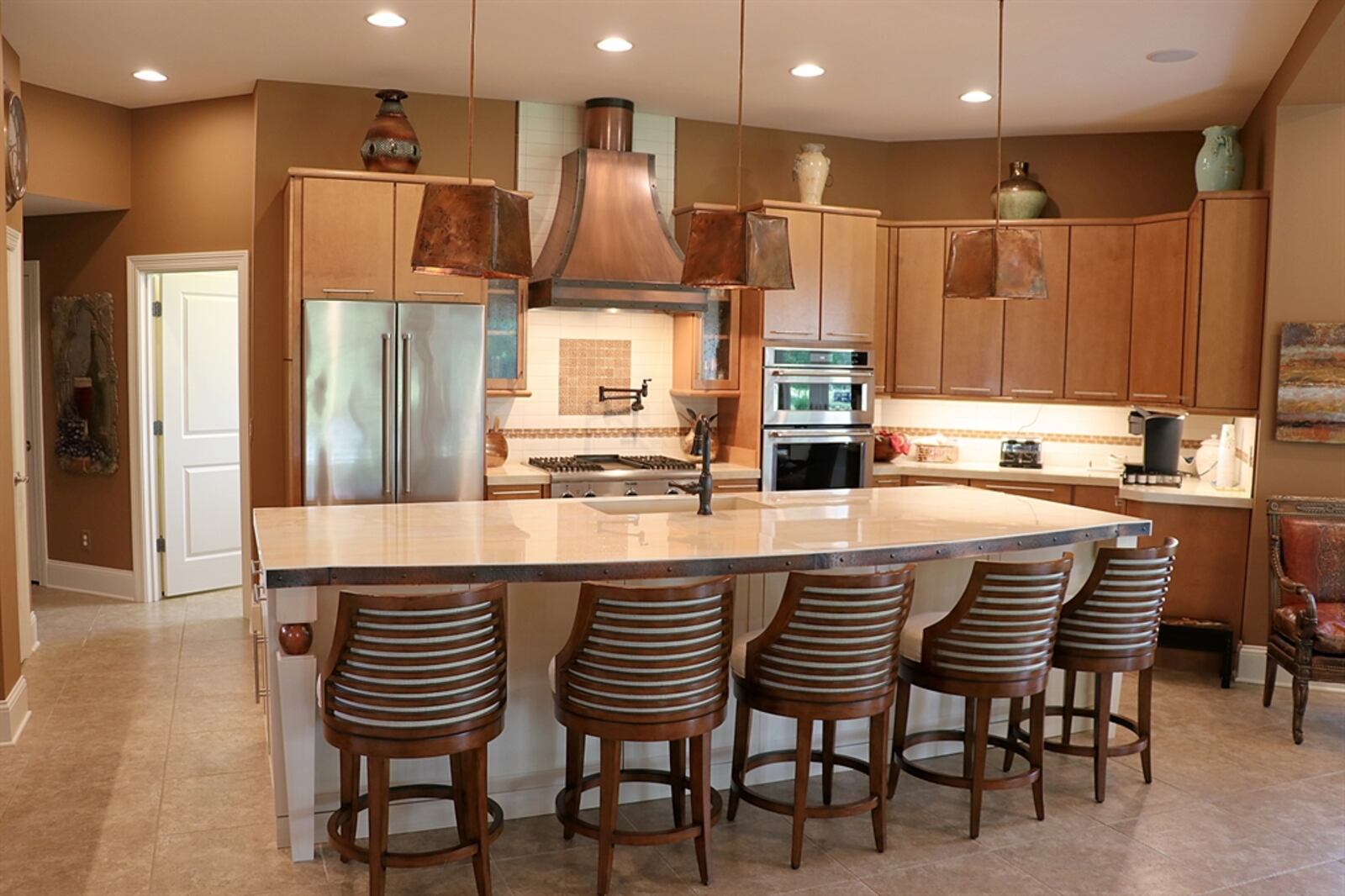 A large island with a cooper accented porcelain countertop extends to offer seating for six as well as hidden storage underneath. Copper bell-shaped lights hang above the island. 