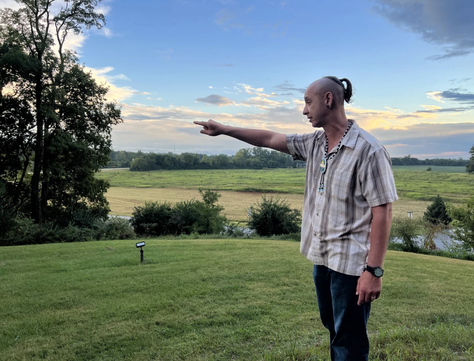 Jeremy Turner (Shawnee Tribe) points to where he thinks a longhouse once stood at a Pe'qa. He is speaking while giving a tour of an 18th-century Shawnee village near modern-day Springfield, Ohio. Fields of row crops can be seen in the background along the Mad River floodplain. Corn has been planted there (first by Shawnee and then Europeans) for hundreds, if not thousands, of years. (Courtesy: Chris Welter, WYSO)