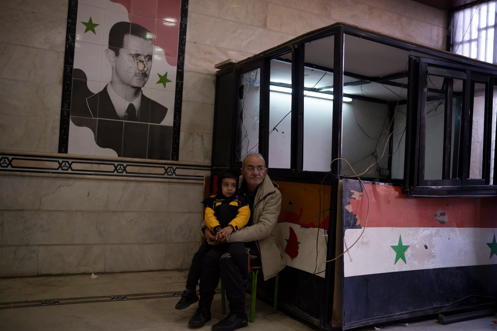 A man sits with a child next to a damaged image depicting the ousted Syrian President Bashar Assad, at the entrance of the Police headquarters, in Homs, Syria, Thursday, Dec. 26, 2024. (AP Photo/Leo Correa)