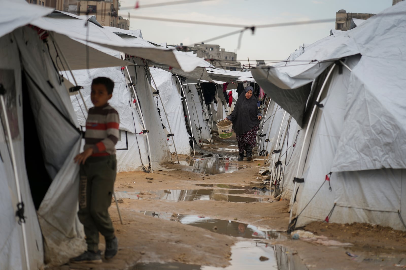 A Palestinian woman carried a muddy bucket as she walks between tents in a sprawling tent camp adjacent to destroyed homes and buildings in Gaza City, Gaza Strip, Saturday, March 1, 2025 during the Muslim holy month of Ramadan. (AP Photo/Abdel Kareem Hana)