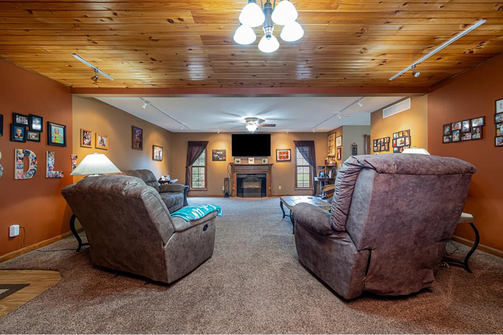 The oversized first level living room has neutral carpeting, wood beaded ceiling (Partial), ceiling fan and recessed lighting. It also has an electric fireplace with wood mantel and surround.