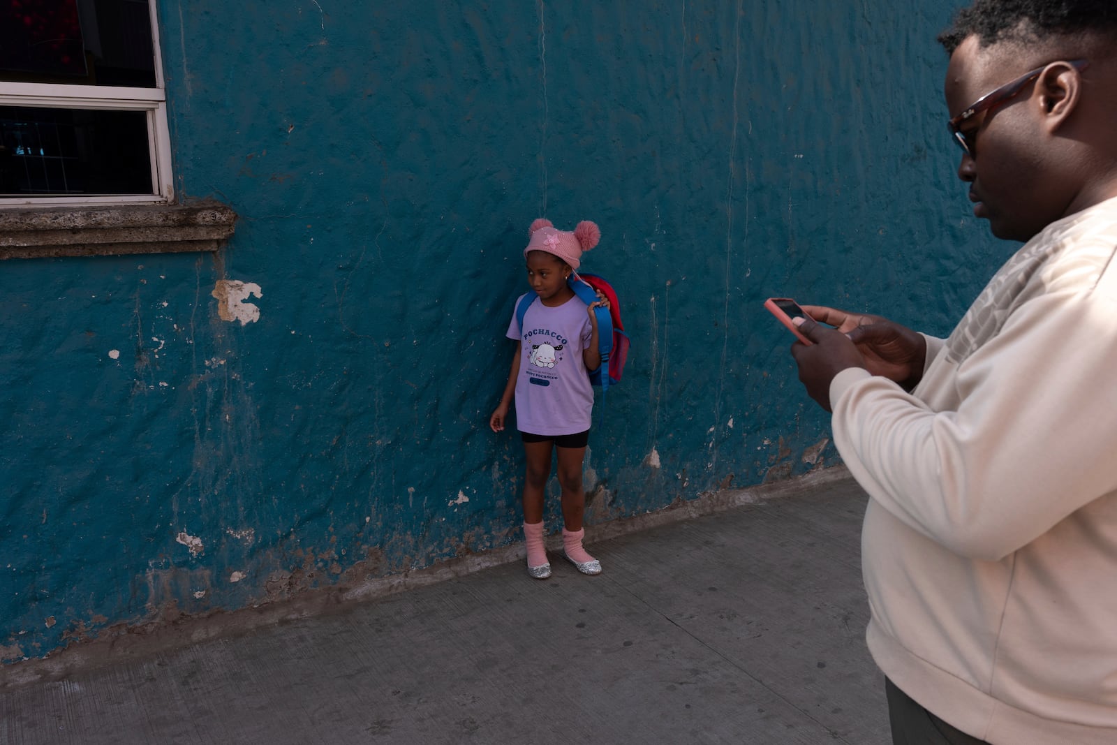 Maickeliys Rodriguez, of Venezuela, 6, wears her newly donated silver slippers as she stops to adjust her backpack under the watchful gaze of a family friend from Haiti, as her family and others from a migrant shelter make their way to a nearby laundromat in Tijuana, Mexico, Friday, Jan. 31, 2025. (AP Photo/Gregory Bull)