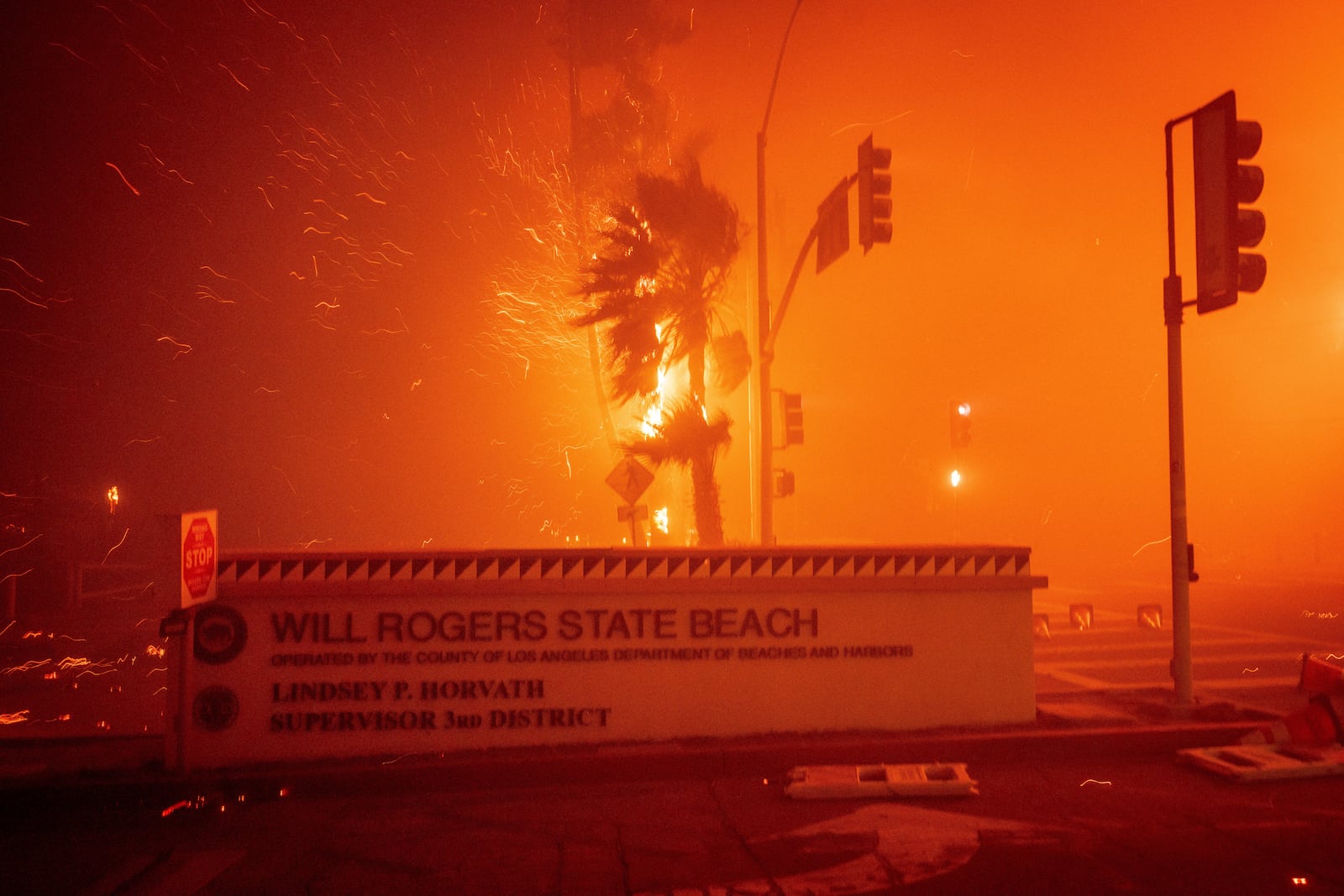 The Palisades Fire burns behind a sign for Will Rogers State Beach in the Pacific Palisades neighborhood of Los Angeles, Tuesday, Jan. 7, 2025. (AP Photo/Ethan Swope)