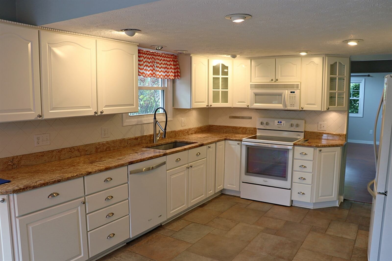 White cabinetry fills the kitchen and surrounds appliances. Granite counters provide plenty of preparation space and a deep sink is below a window. Pantry cabinets surround the refrigerator, and the corner cabinets have glass-panel doors.