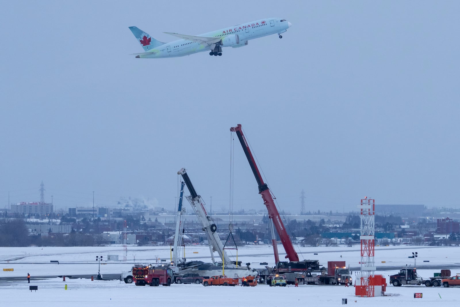 An Air Canada flight takes off in the background as cranes are positioned to remove the wreckage of Delta Flight 4819 from the runway at Toronto Pearson International Airport, in Mississauga, Ontario, on Wednesday, Feb. 19, 2025. (Arlyn McAdorey/The Canadian Press via AP)