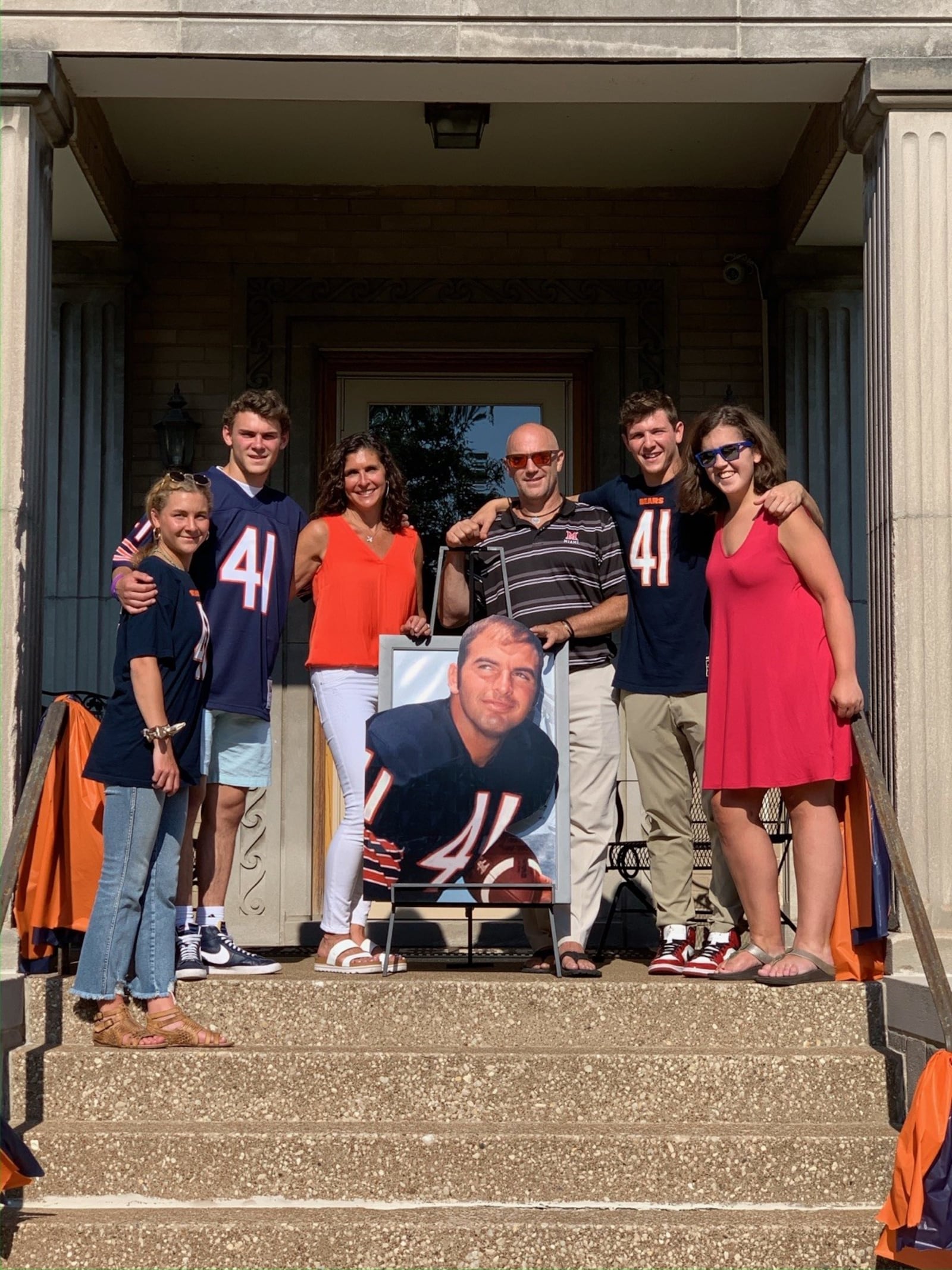 Two years ago – on the 50th anniversary of Brian Piccolo’s death – a parade was held in Beverly, the far south side community in Chicago where Piccolo, his wife Joy and their three young daughters lived wehn he played for the Chicago Bears. Although many of the festivities planned by the Bears were cancelled by COVID-19 concerns, the grassroots parade drew over 40 cars and went from Piccolo’s Christ the King Catholic Church to the two home where his family once lived. Joining the festivities that day was the family of Piccolo’s middle daughter, Traci Piccolo Dolby: (from left to right) Traci’s daughter Drew; her son Brian, a walk-on wide receiver for the Dayton  Flyers who transferred from Colorado State; her husband John, holding the portrait of Piccolo; son Jack and daughter Grace. CONTRIBUTED