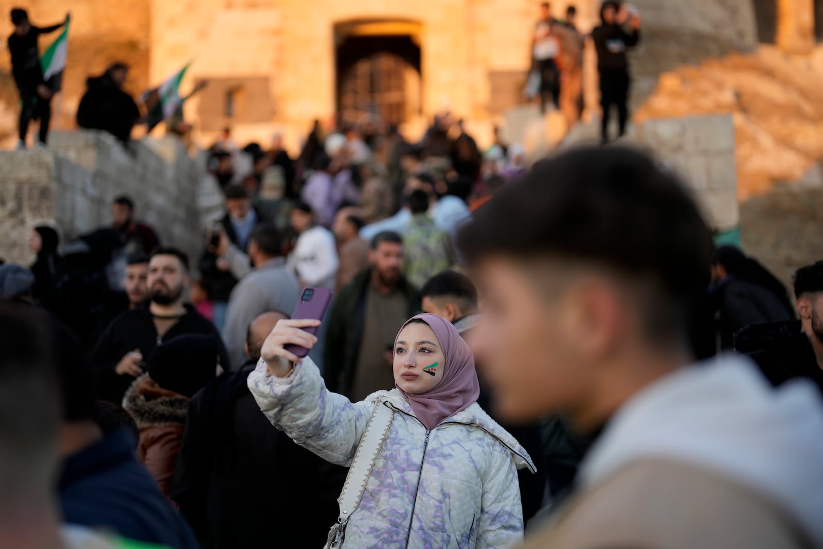 A Syrian girl with the colours of the "revolutionary" Syrian flag on her face, takes a selfie in front of the ancient Aleppo Citadel in the old city of Aleppo, Syria, Saturday, Dec. 14, 2024. (AP Photo/Khalil Hamra)